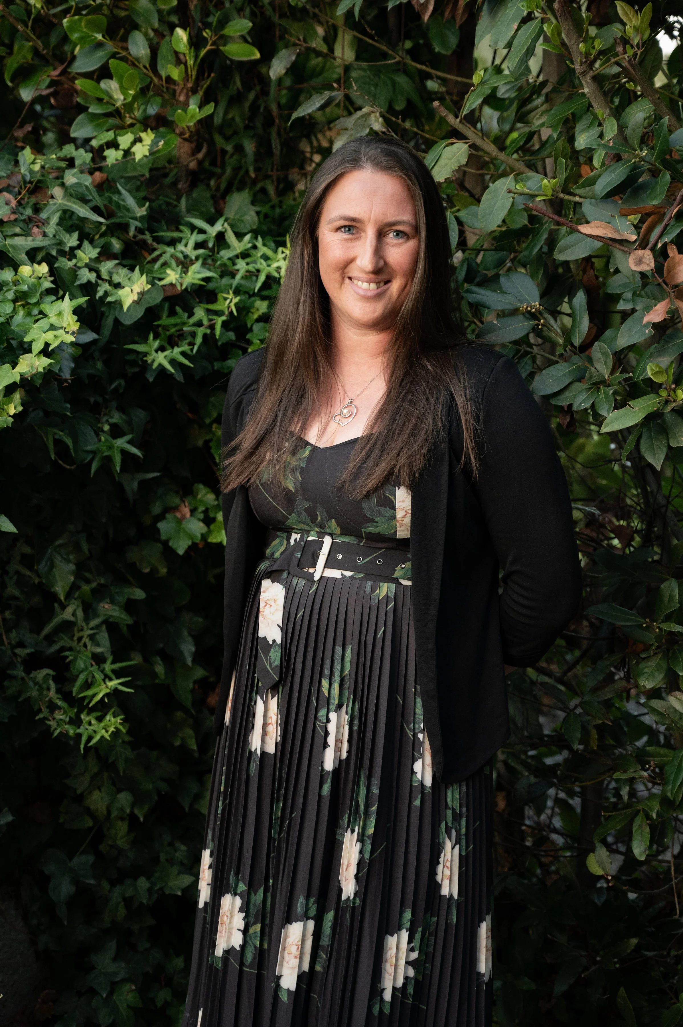 A woman with long brown hair and a black dress with floral patterns, standing in front of green foliage, smiling at the camera.
