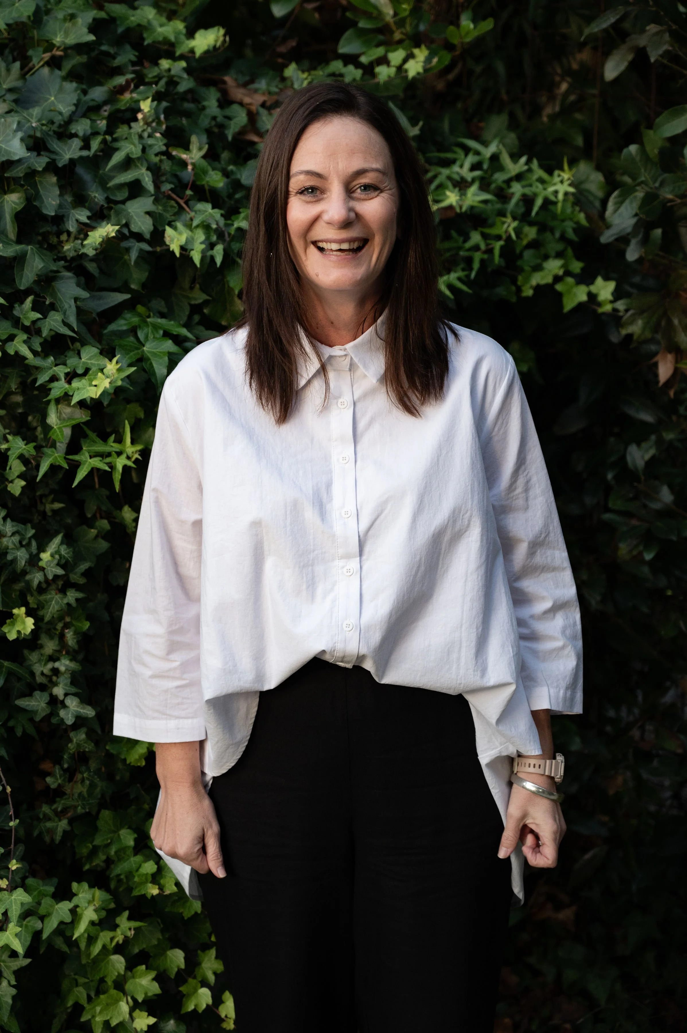 A woman with long brown hair, smiling, wearing a white, loose-fitting button-up shirt and black pants, standing outdoors in front of green leafy plants.