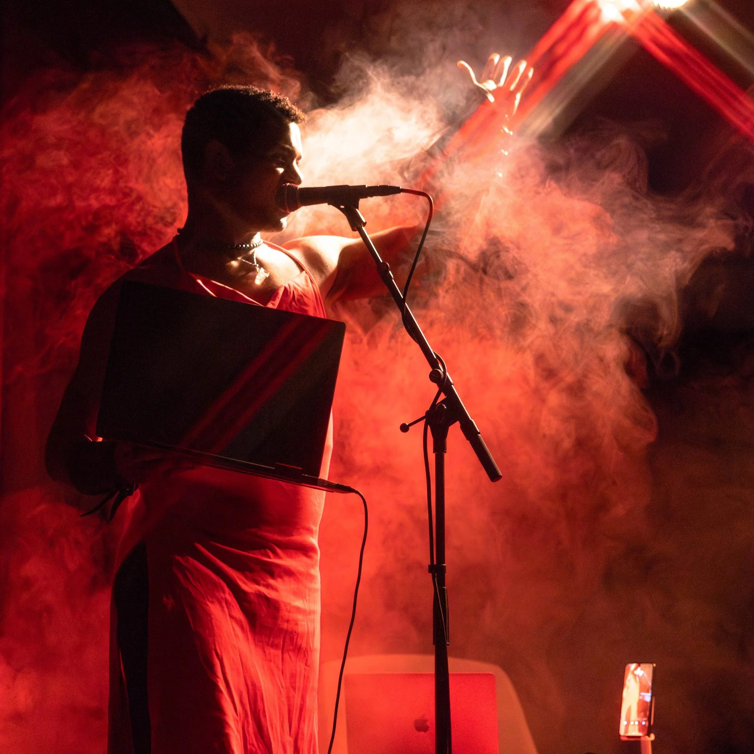 Rohan Zhou-Lee in a red dress and smoky room speaking into a microphone. Photo credit Lehan Zhang