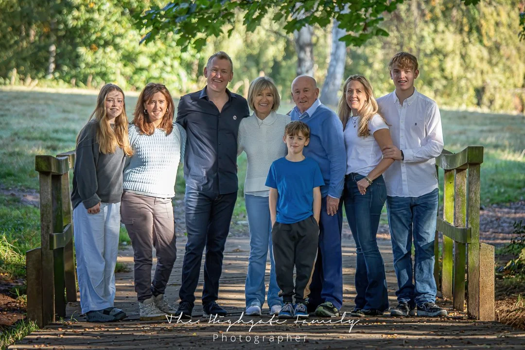A large family of nine people standing on a wooden bridge outdoors, smiling, with green trees and grass in the background.