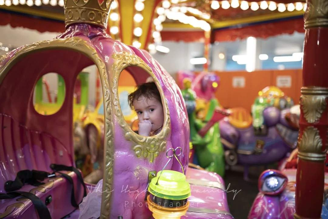 Young girl with curly hair sitting inside a pink and gold carousel ride at an amusement park, looking at the camera with her hand near her mouth.