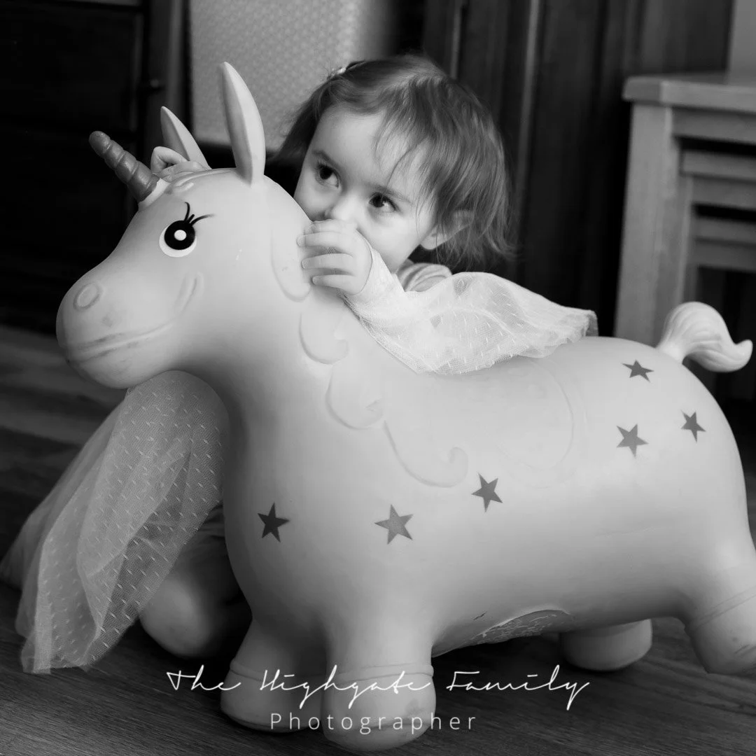 A young girl with curly hair playing with a large inflatable unicorn toy, looking over its head and hugging its horn, in a black and white photo.