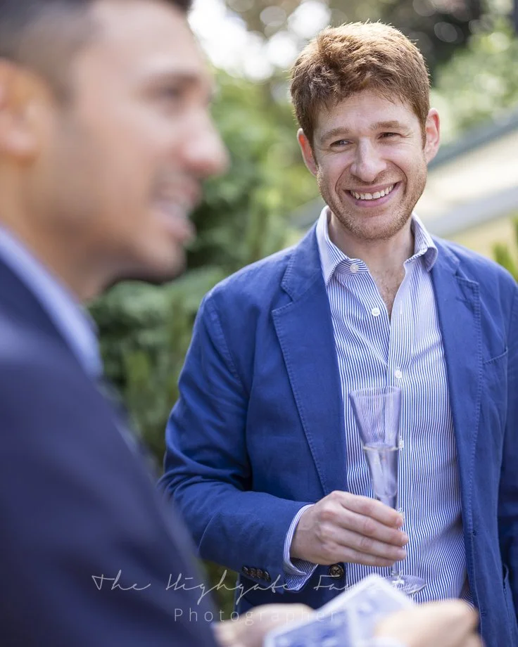 Two men in formal attire at an outdoor event, one smiling and holding a champagne flute, the other blurred in the foreground.