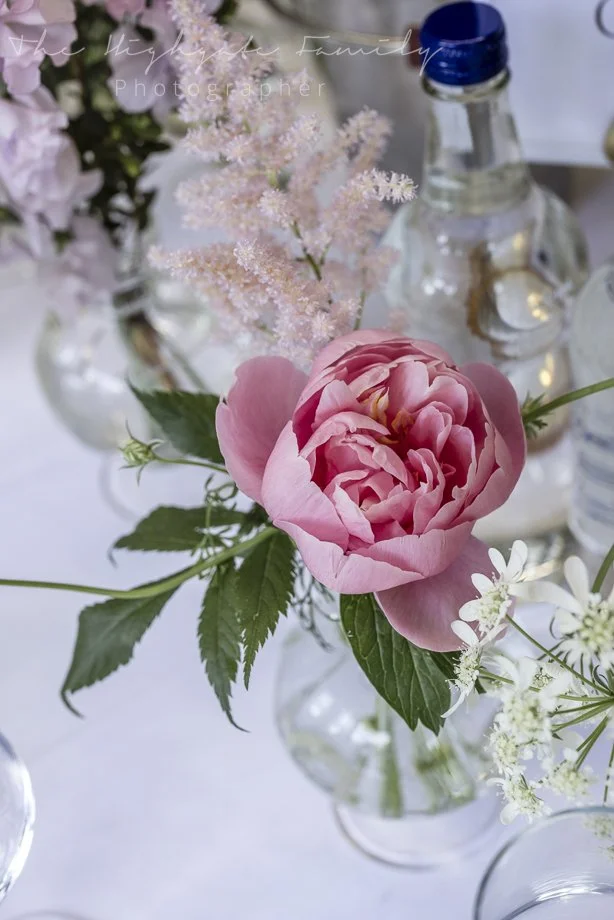 Close-up of a pink peony flower in a glass vase surrounded by green leaves, with other floral arrangements and bottles in the background.