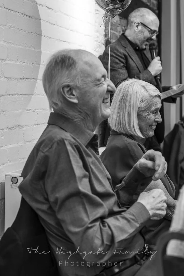 A black and white photo of a smiling elderly man with short hair and a woman with shoulder-length hair, sitting next to each other. A man in the background is speaking into a microphone. There is a balloon near the speaker and a brick wall behind the