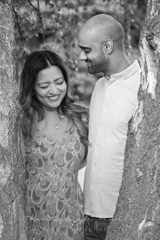A smiling woman and a man stand between two trees, looking at each other in a park or outdoor setting.
