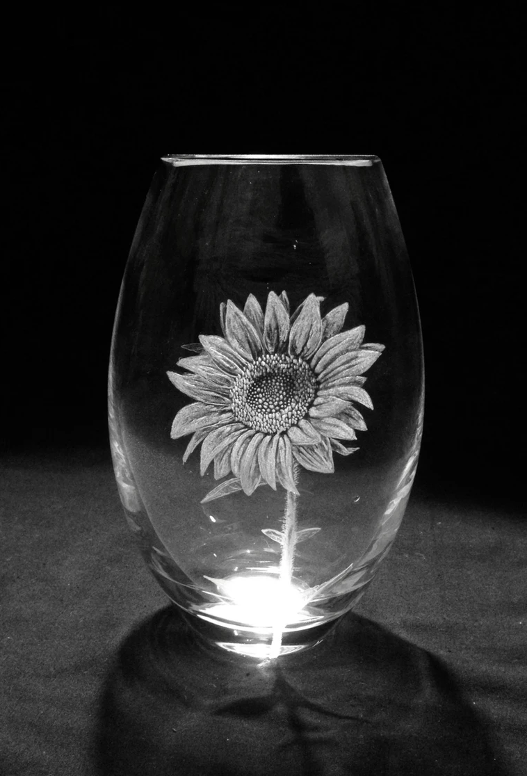 Vase en verre transparent avec une peinture d'un tournesol, éclairé par une source de lumière en dessous, sur un fond noir.