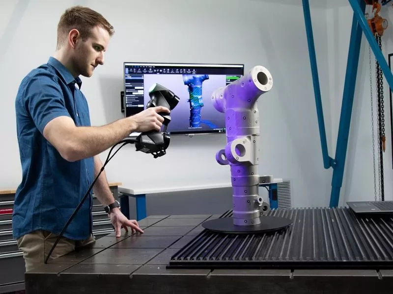 Un homme utilise un scanner 3D pour analyser un bras robotique sur une table de travail, avec un écran à l'arrière affichant une modélisation numérique du bras.