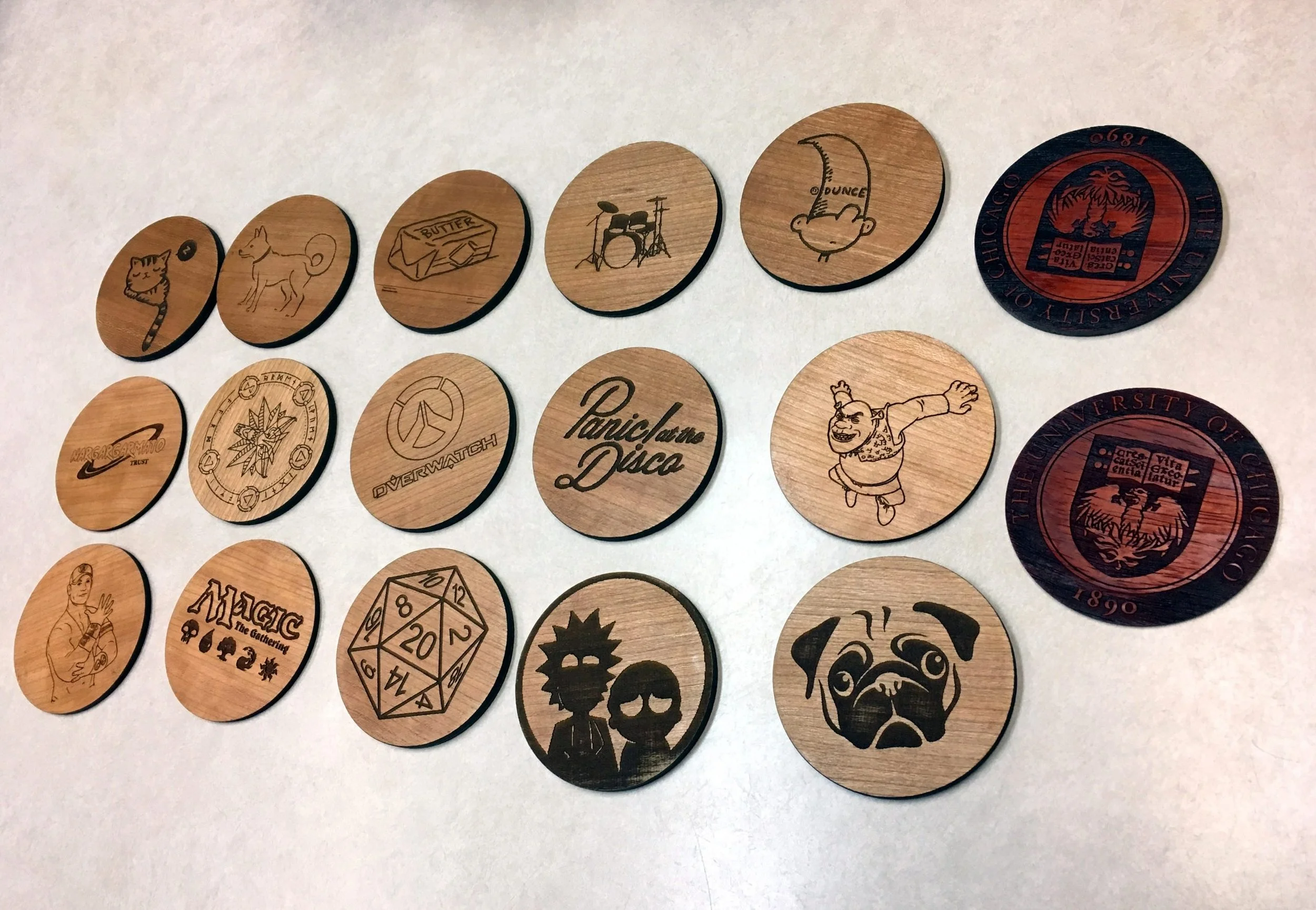Trois rangées de 6 pièces de bois avec divers dessins et textes gravés, comprenant un chat, un chien, du beurre, une batterie, un clown, le logo d'Overwatch, un dessin de drapeau, une femme, un rouleau de parchemin, un personnage animé, un visage de chien, et d'autres images variées.