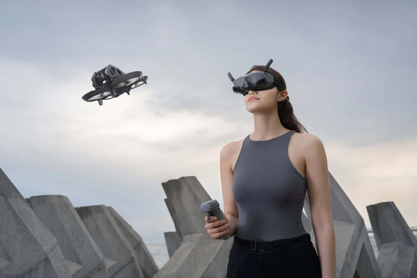 Une femme portant des lunettes de réalité virtuelle tient une manette, avec un drone volant à proximité, sur un quai au bord de l'eau, avec un ciel nuageux en arrière-plan.