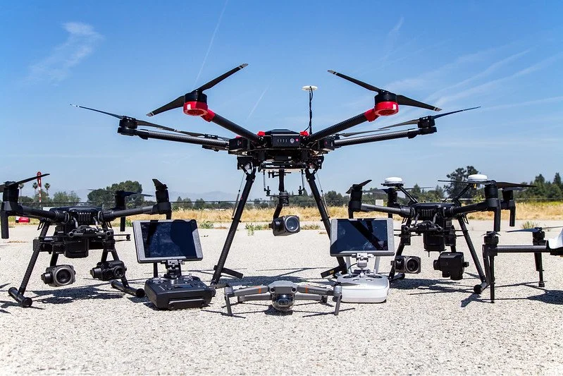 Trois drones de différents modèles posés sur un sol extérieur, équipés de caméras et écrans, avec un ciel bleu en fond.