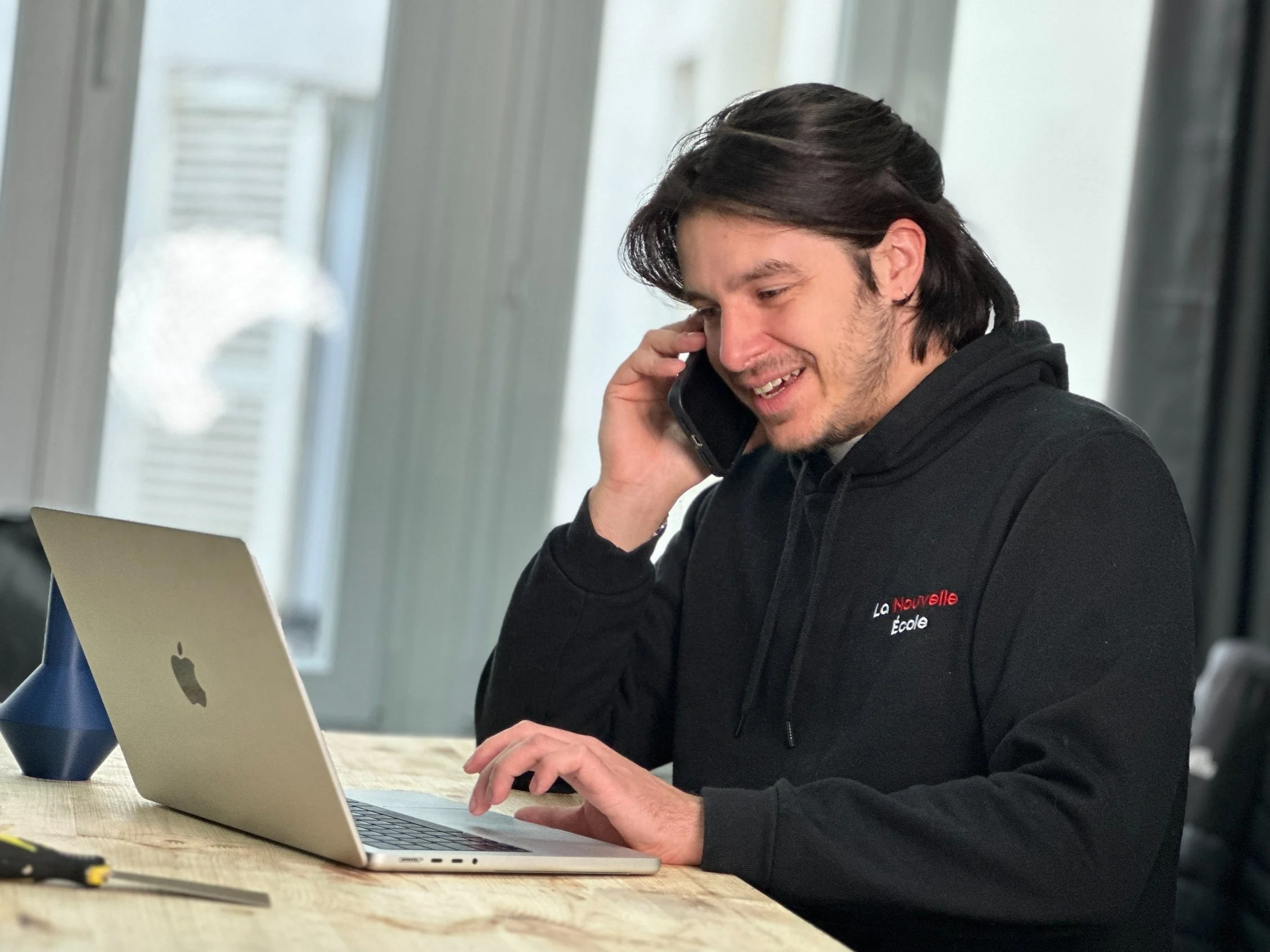 Jeune homme avec long cheveux noirs, souriant, portant un hoodie noir avec l'inscription "La Nouvelle École", utilisant un ordinateur portable sur une table en bois, tout en parlant dans un téléphone portable. Fonds lumineux avec des fenêtres.