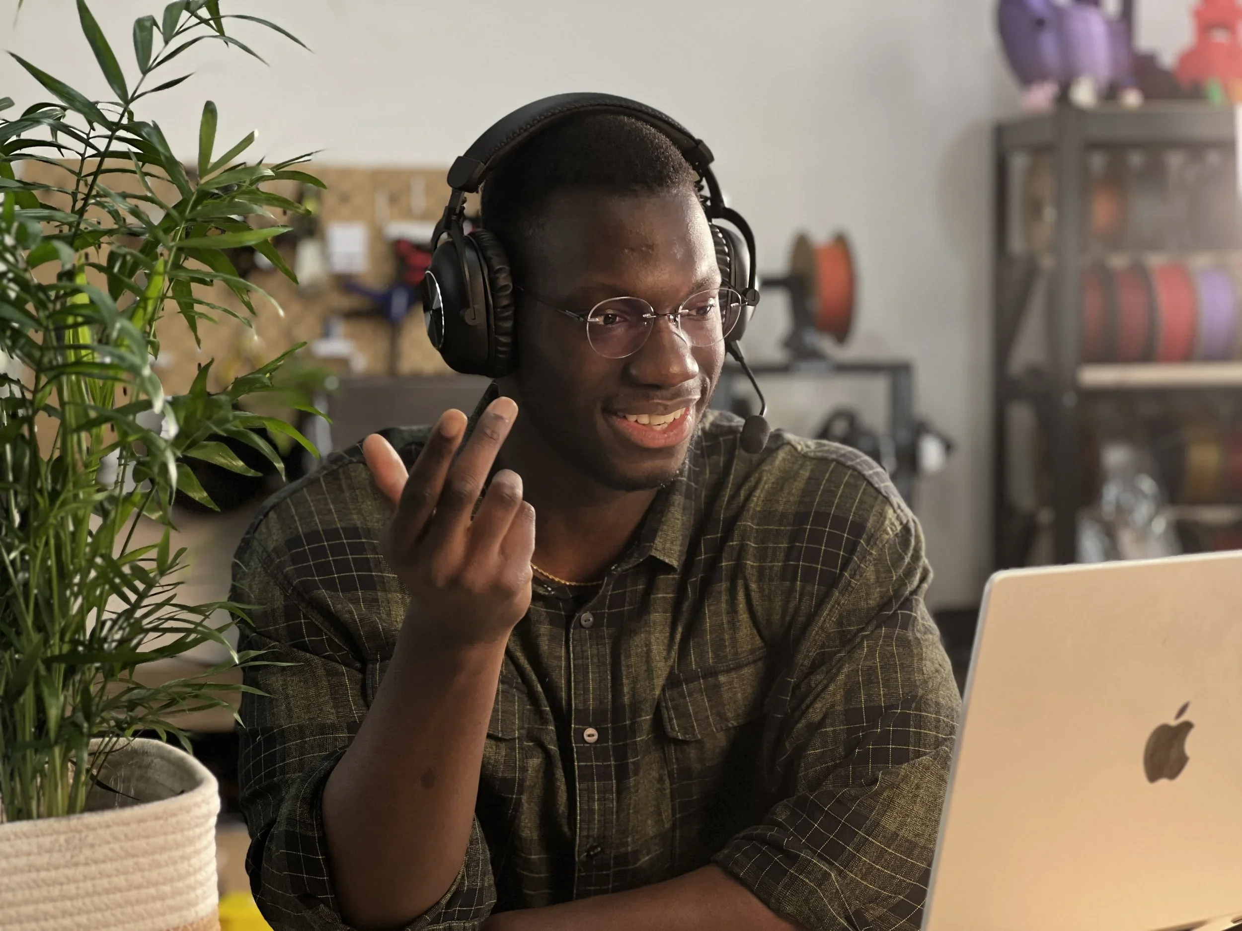 Un jeune homme afro-américain portant un casque d'écoute et des lunettes, assis devant un ordinateur portable Apple, dans un espace intérieur avec des plantes et des meubles en arrière-plan.