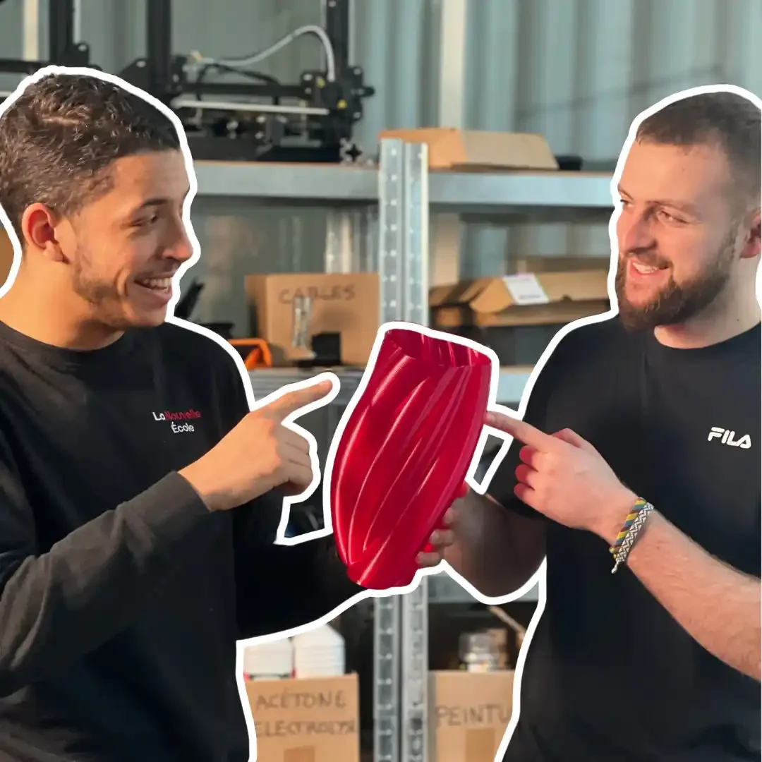 Deux hommes de front s'amusent, pointant du doigt un gros objet rouge entre eux, dans un atelier ou un espace de stockage.