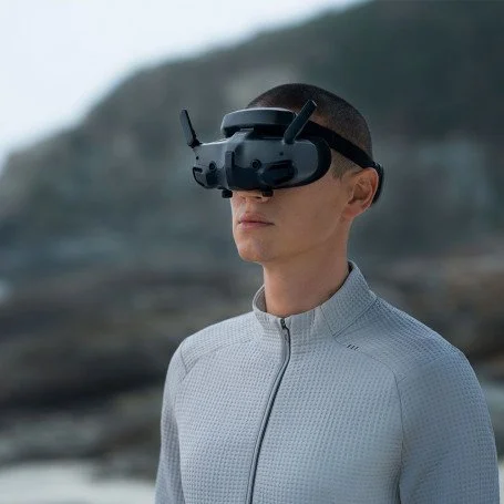 Une personne portant un casque de réalité virtuelle avec des antennes, en extérieur avec un paysage naturel en arrière-plan.