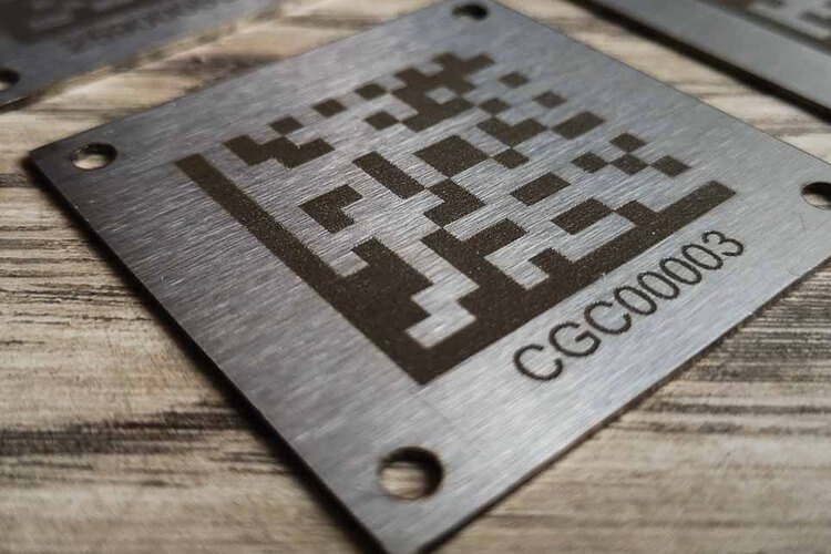 Plaque en métal avec un motif QR code gravé, posée sur une surface en bois.