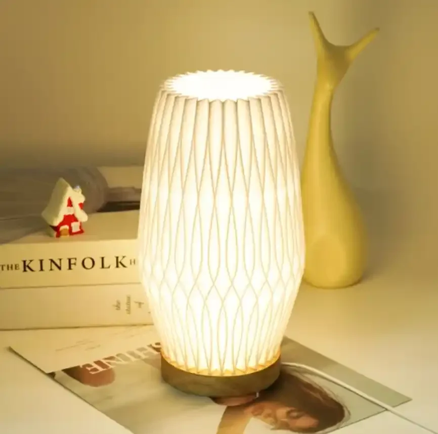 Lampe de table allumée avec un abat-jour en papier blanc, posée sur un magazine avec une personne sur la couverture, accompagnée d'une petite figurine blanche et rouge, d'un livre intitulé 'The Kinfolk' et d'un vase jaune.