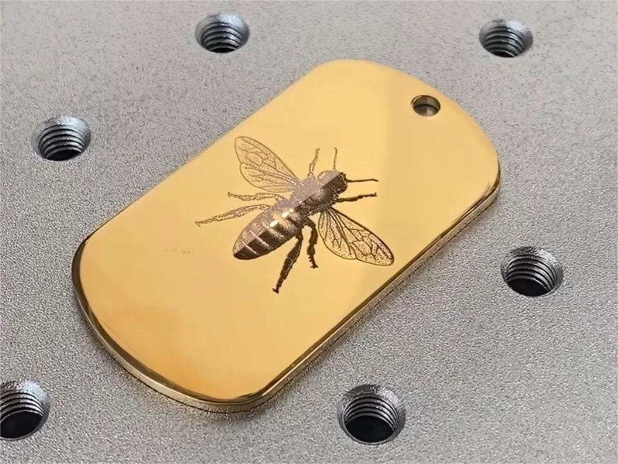 Une plaque métallique dorée avec une image d'une abeille gravée, posée sur une surface grise avec plusieurs trous filetés noirs.