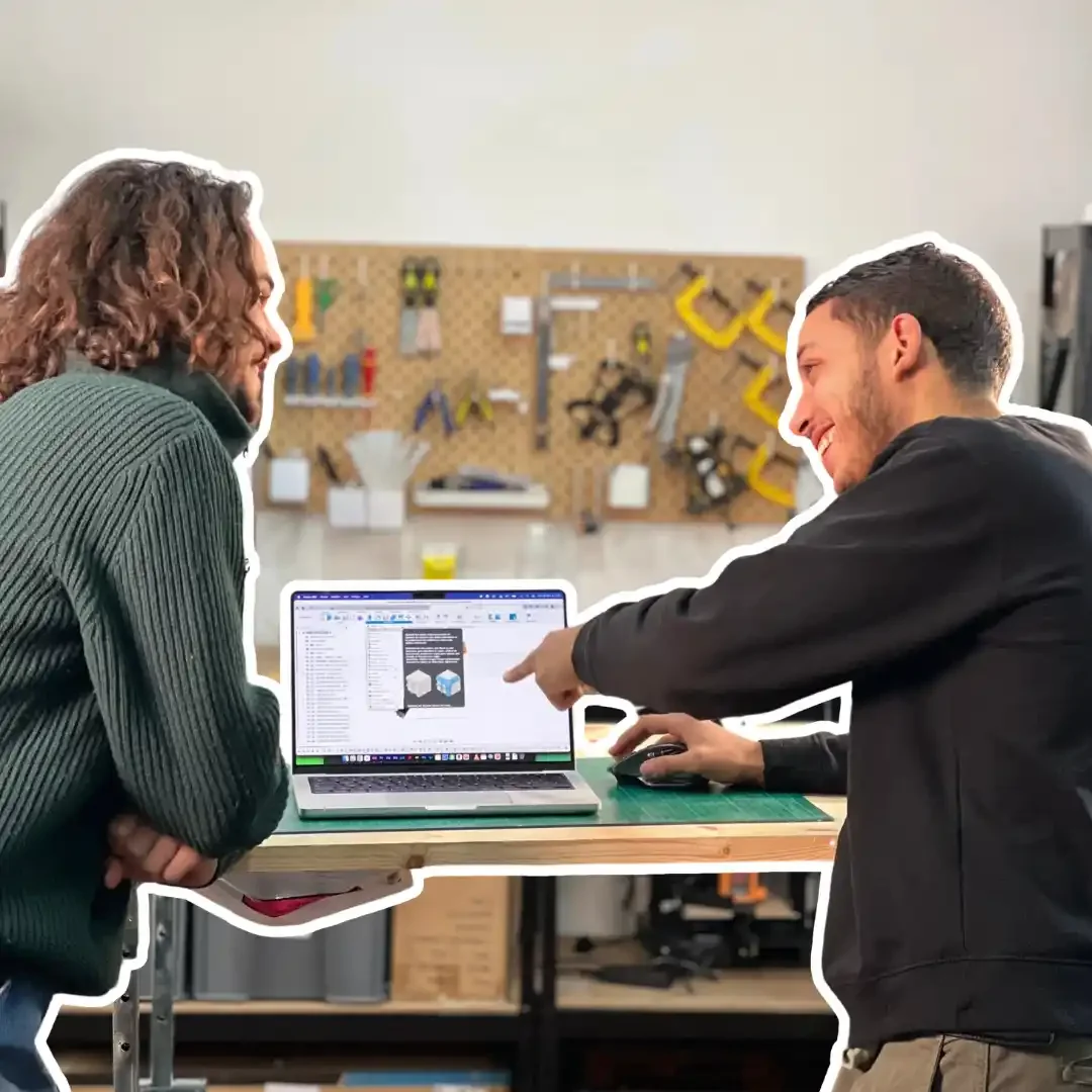 Deux hommes discutent devant un ordinateur portable sur un bureau en bois dans un atelier, un des hommes pointe l'écran. En arrière-plan, un panneau de bricolage avec des outils suspendus.
