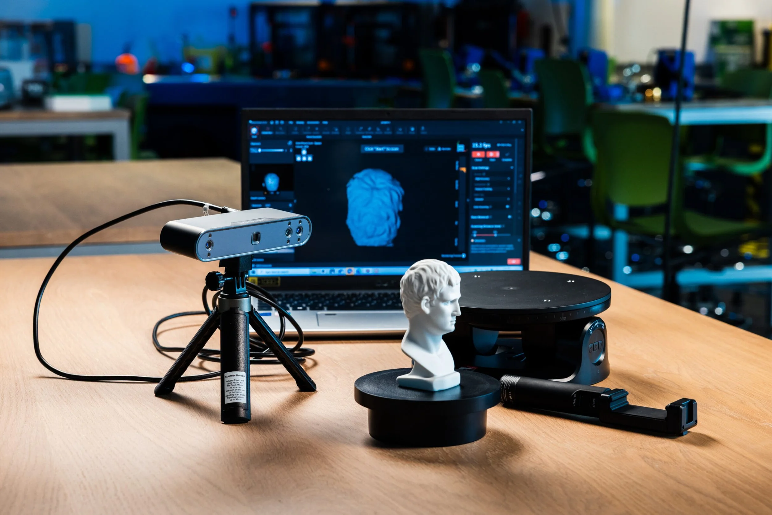 Une table de travail avec un ordinateur portable affichant une modélisation 3D d'un visage, un scanner 3D, une tête de sculpture en plâtre, et d'autres équipements de numérisation 3D dans un laboratoire ou atelier.