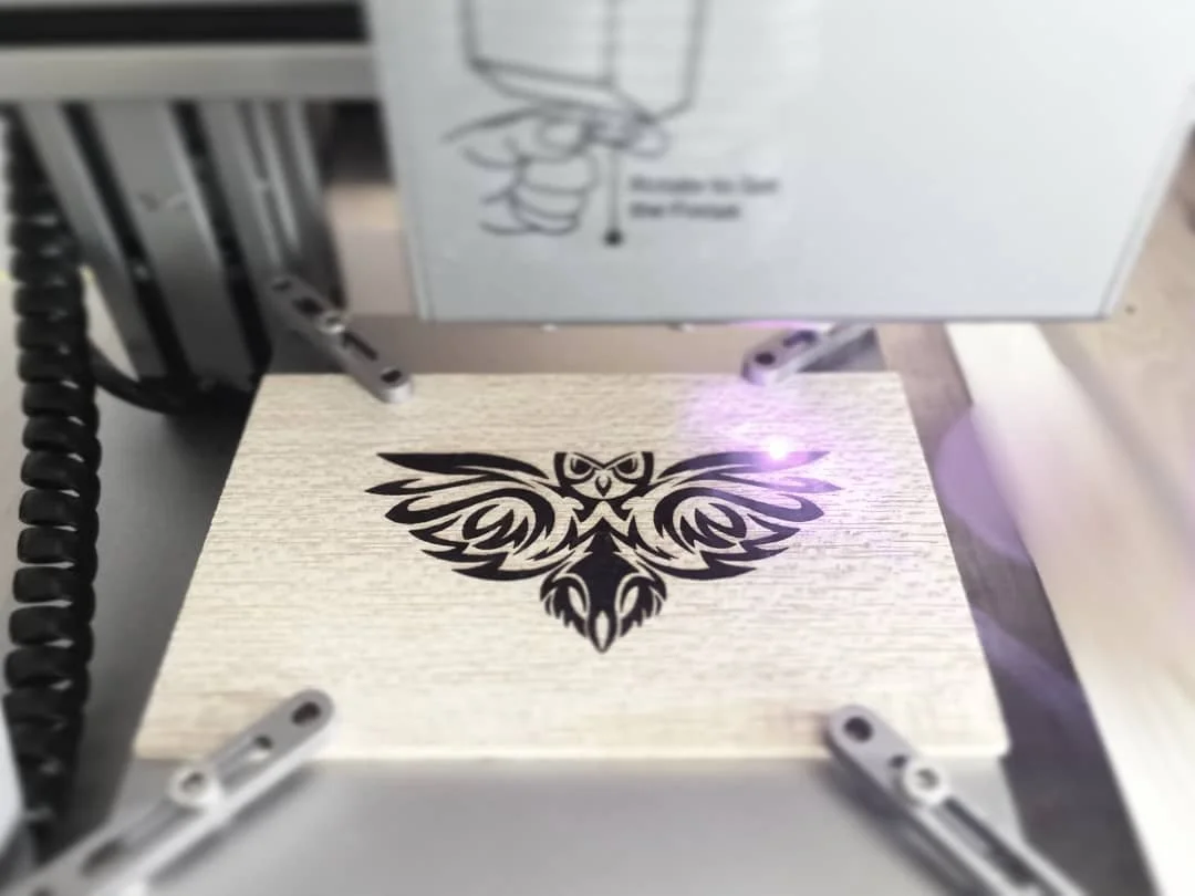 Une machine de gravure laser inscrivant une victoire avec un hibou stylisé sur une plaque en bois.