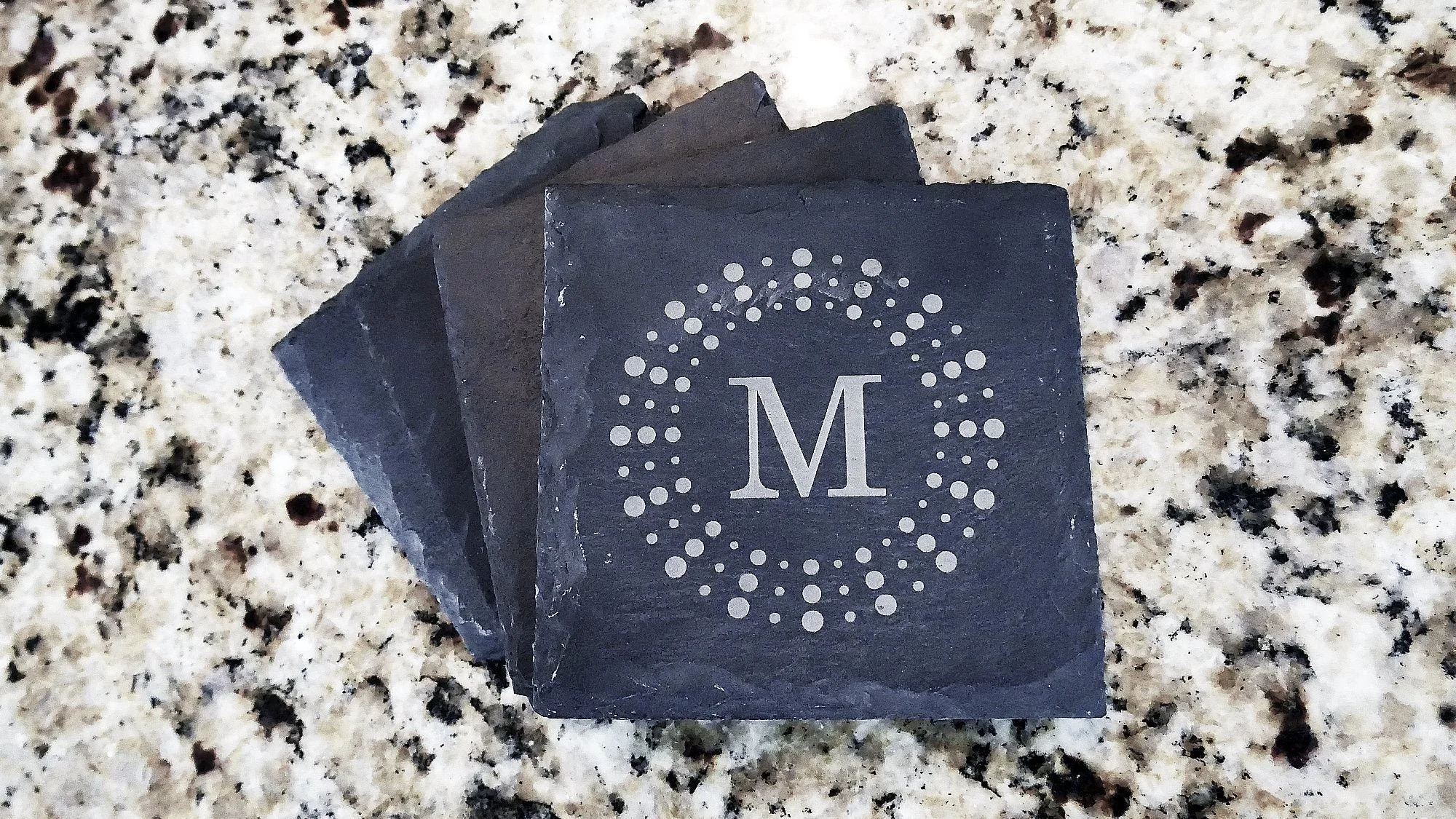 Dessus de tuiles en ardoise gravé avec un cercle de points et la lettre M au centre, posé sur un plan de travail en granite.