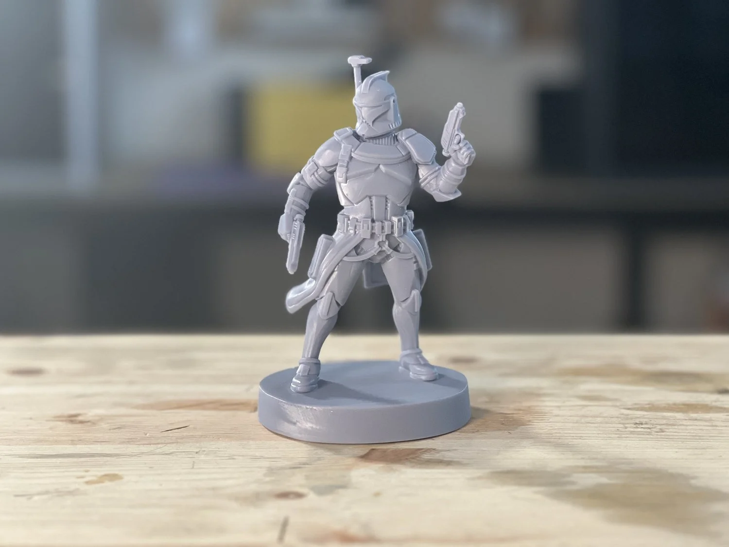 Une figurine de personnage de l'univers Star Wars, semblable à un chasseur de primes ou un soldat, en plastique gris clair, avec un casque et un pistolet, positionnée sur une base ronde en plastique, sur une table en bois.