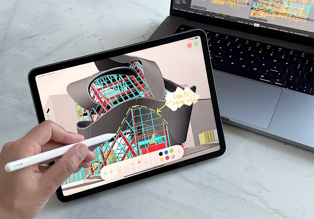 Une personne utilise une tablette pour faire une annotation d'une illustration d'un bâtiment avec des détails en couleur, tandis qu'un ordinateur portable avec un écran montrant un logiciel de conception est visible à côté.
