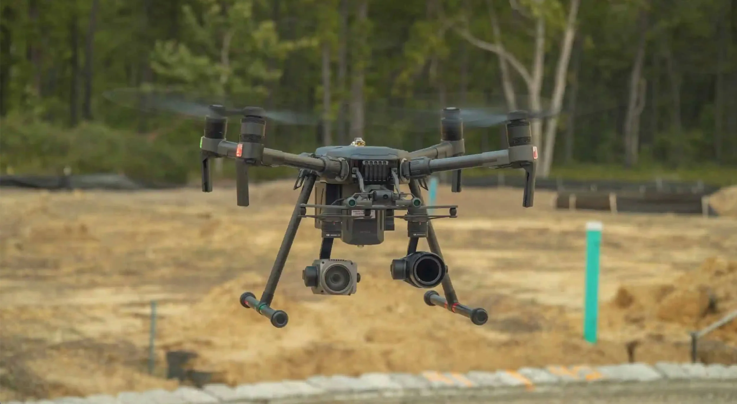 Un drone de surveillance avec plusieurs caméras, volant au-dessus d'un site en extérieur avec un fond d'arbres et de terre.