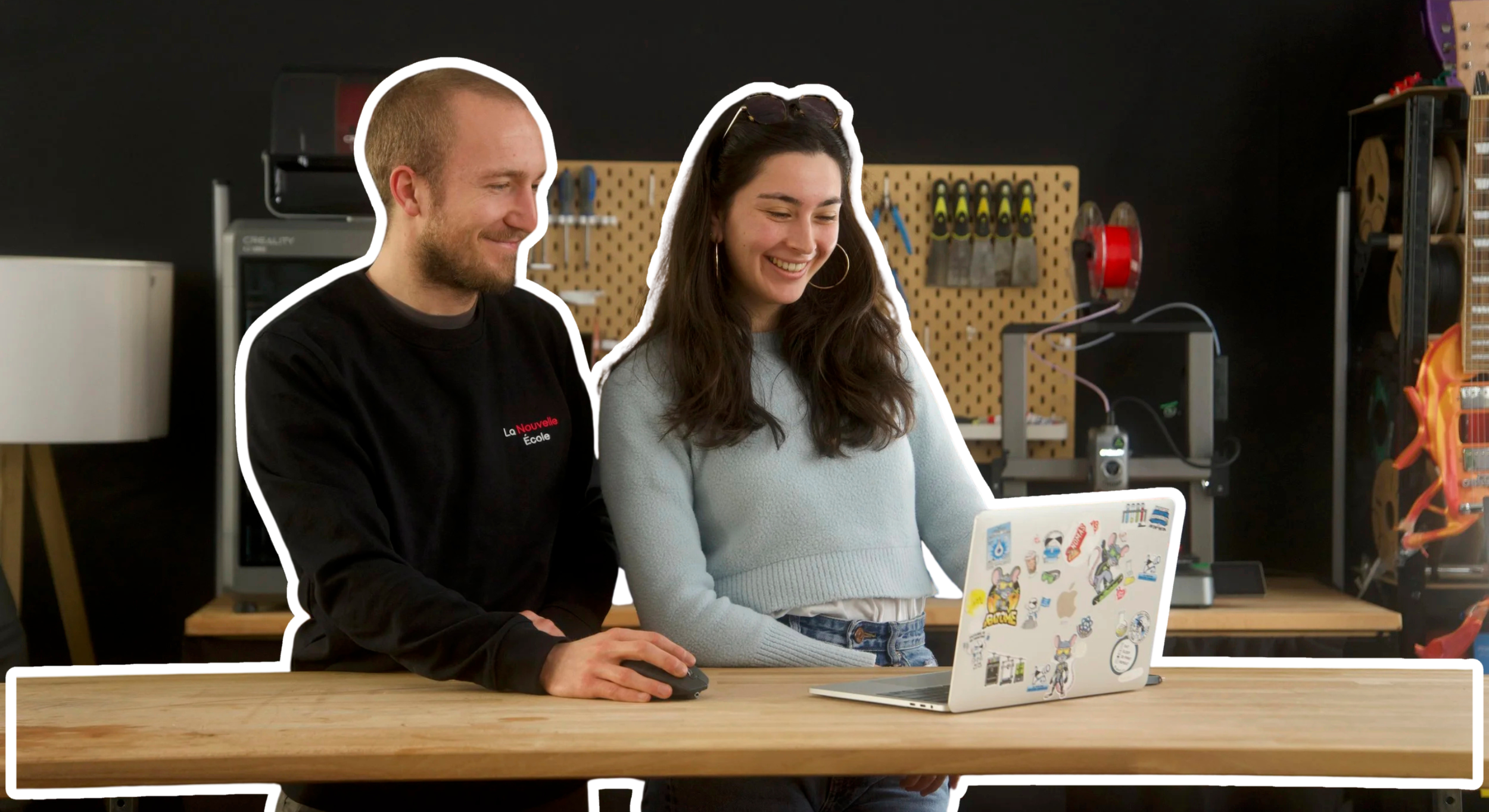 Deux personnes, un homme et une femme, regardent un ordinateur portable décoré de stickers, dans un atelier ou un espace de bricolage.
