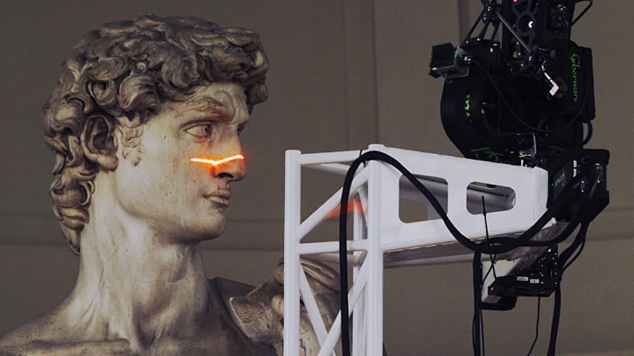 Une sculpture de bust de bronze, représentant un homme avec des cheveux bouclés, une lumière laser rouge traverse son visage, et une caméra ou un dispositif technologique est placé devant la sculpture.