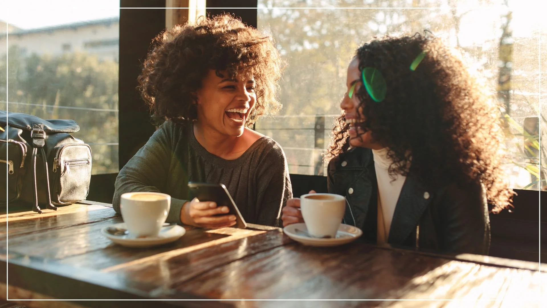 two woman laughing at something on phone while having coffee