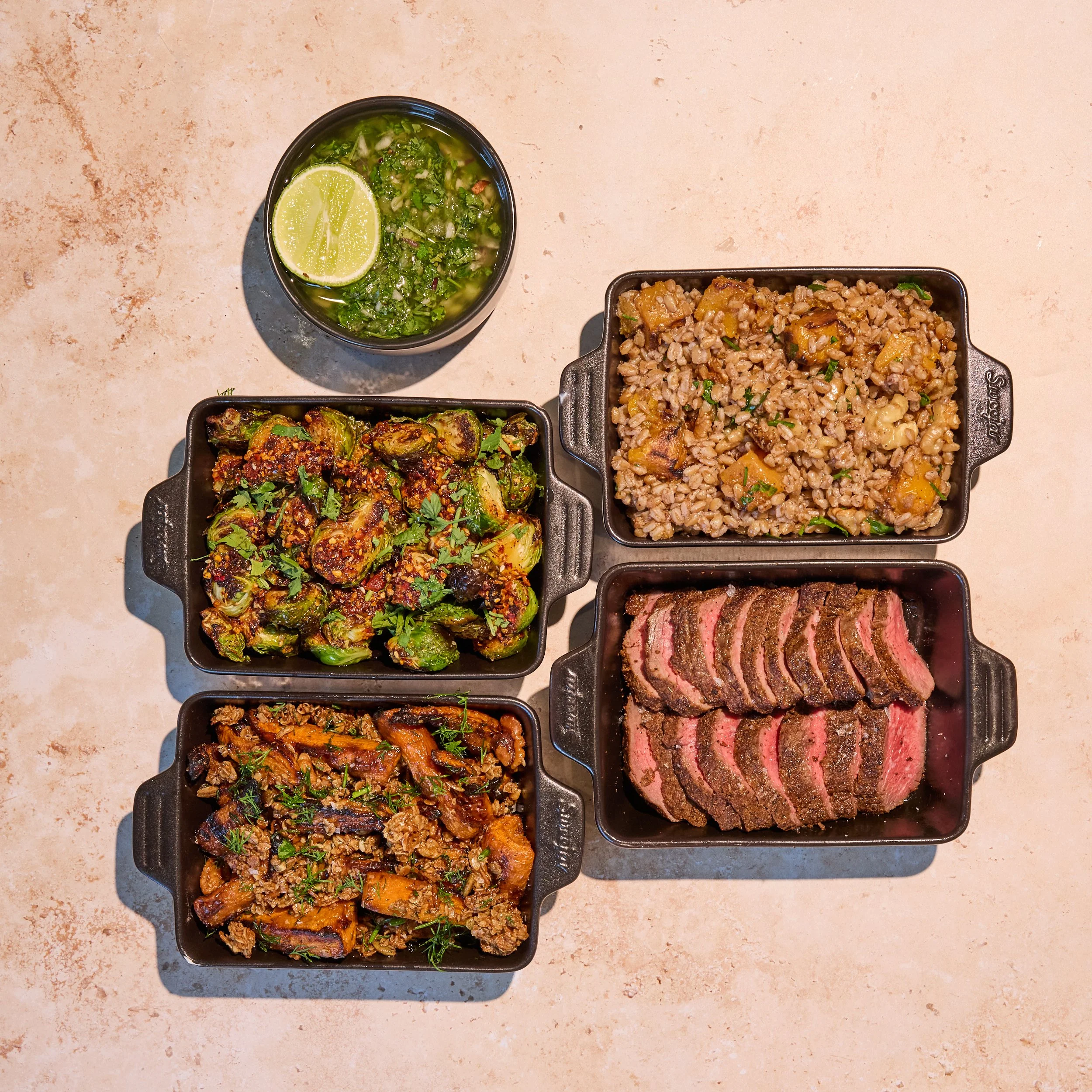 Steak Harvest Family Meal. Overhead view of a restaurant meal with sliced medium-rare steak, roasted Brussels sprouts, glazed carrots, barley and roasted squash, plus chimichurri sauce with lime.