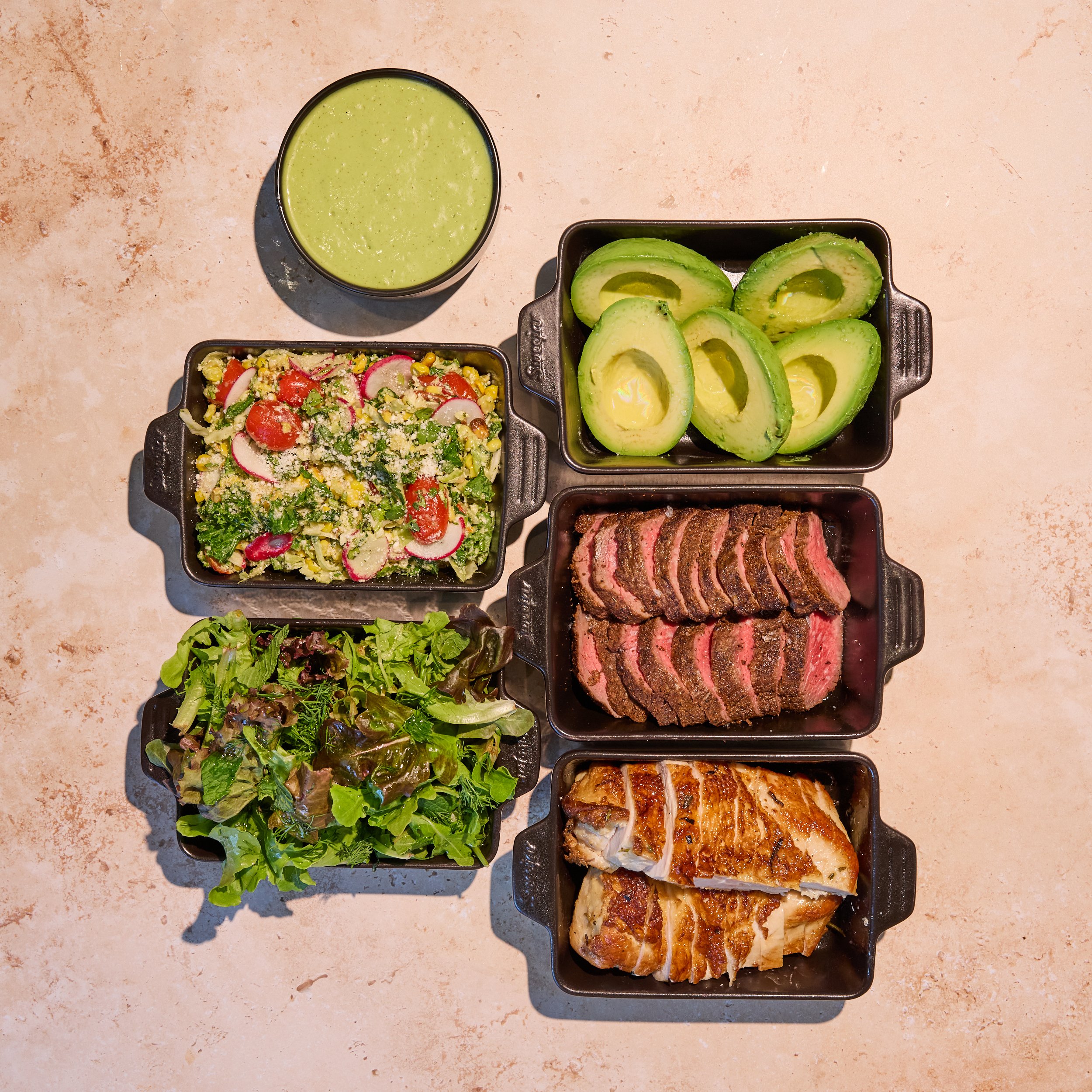 Skinny Protein Family Meal. Overhead spread of sliced steak, roasted chicken, avocado halves, mixed salads, vegetables, and green sauce in black serving dishes.