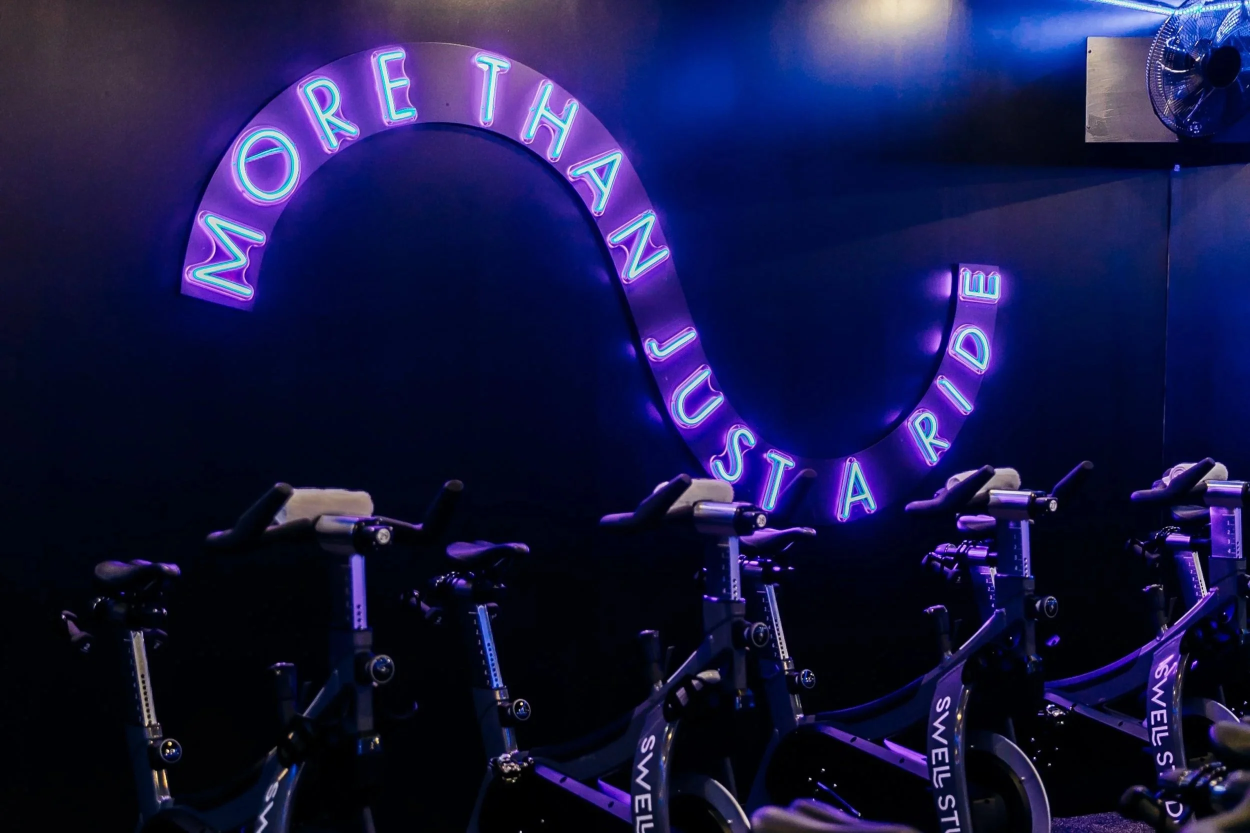 Neon sign in purple and blue reading "More Than Just A Ride" on a dark wall, with several stationary exercise bikes in front of it.