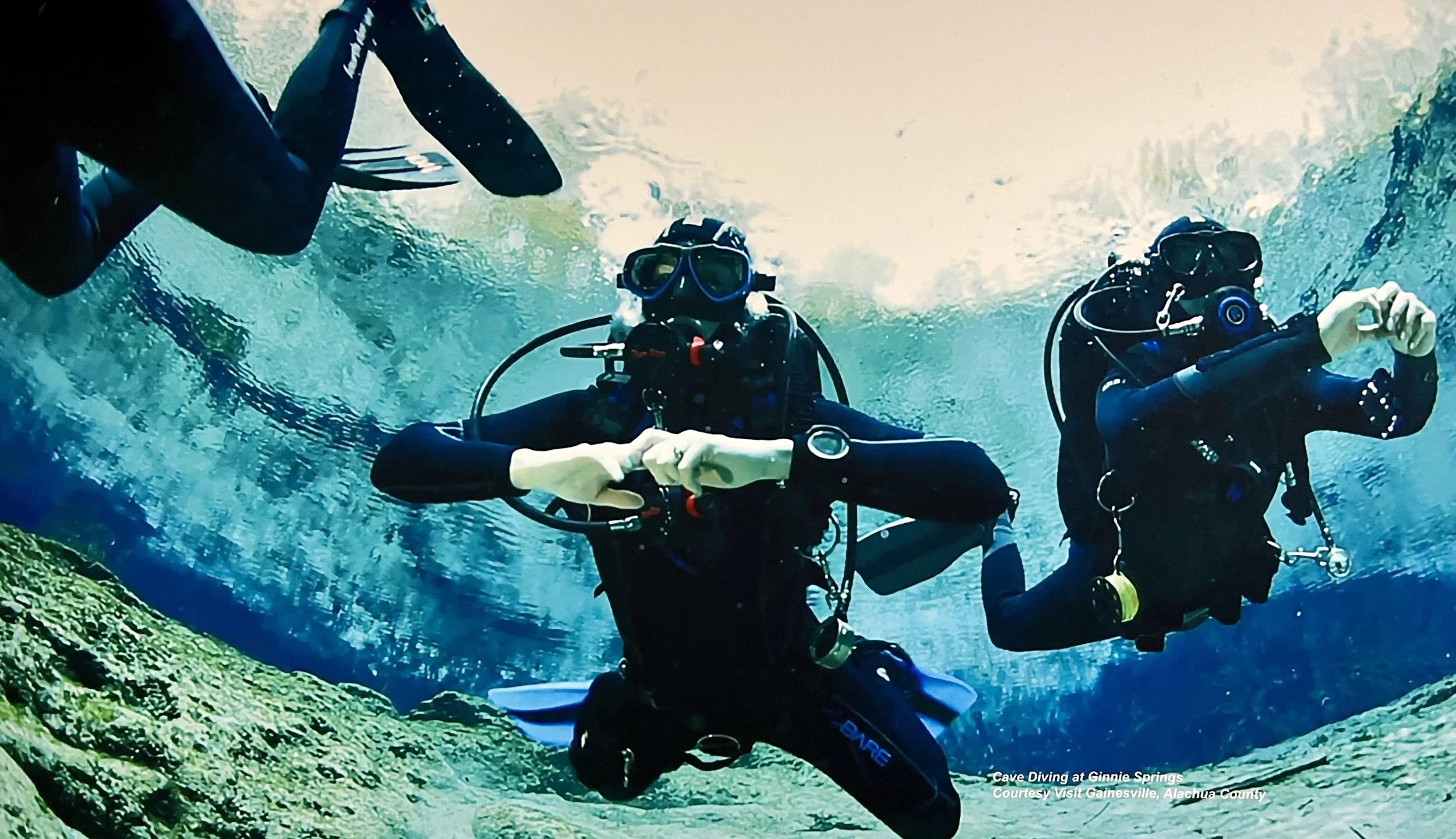 Two scuba divers underwater near a rocky ledge, wearing full diving gear including wetsuits, masks, and tanks, making hand signals.