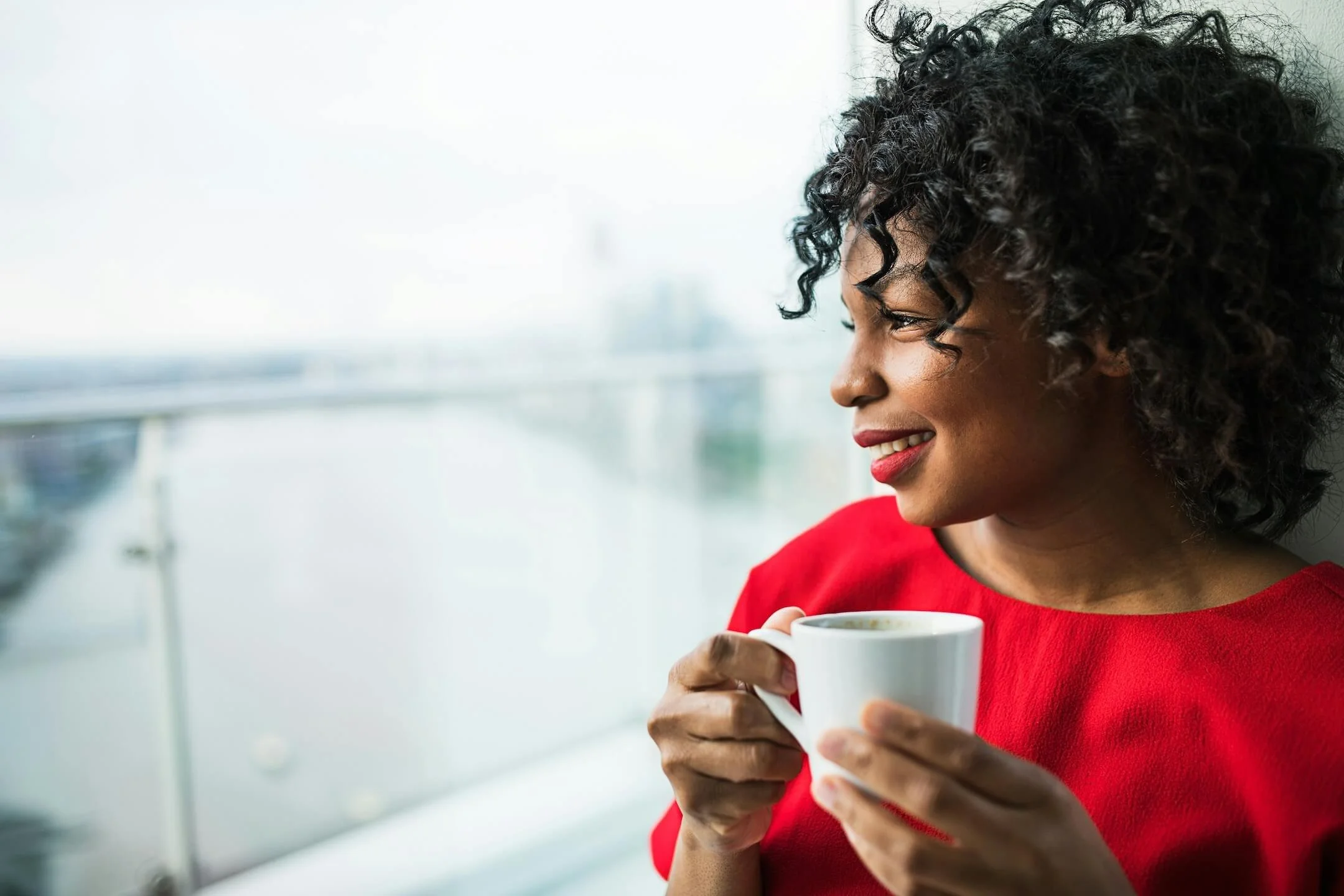 A black woman with curly hair, wearing red lipstick and a red shirt, holds a white coffee cup and smiles while looking out a window EMDR therapy intensives near Boston heal unhealthy relationship patterns