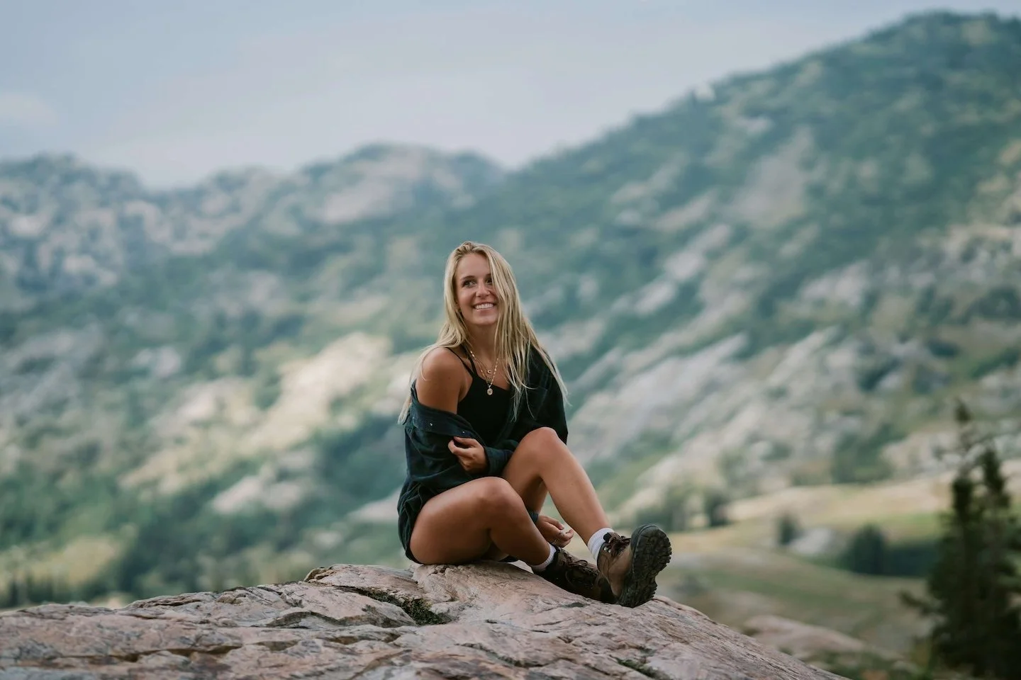 woman with long, blonde hair, wearing shorts, hiking boots, and a flannel shirt, sits smiling on top of a mountain, EMDR healing retreat near Boston