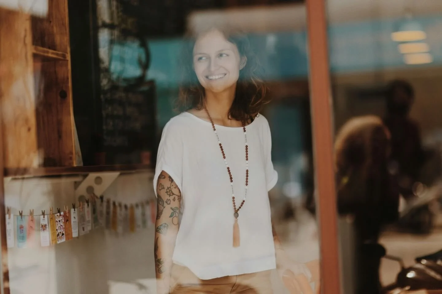 woman standing, looking through a window smiling. She wears a white top with a long, beaded necklace, and has colorful tattoos along her arm