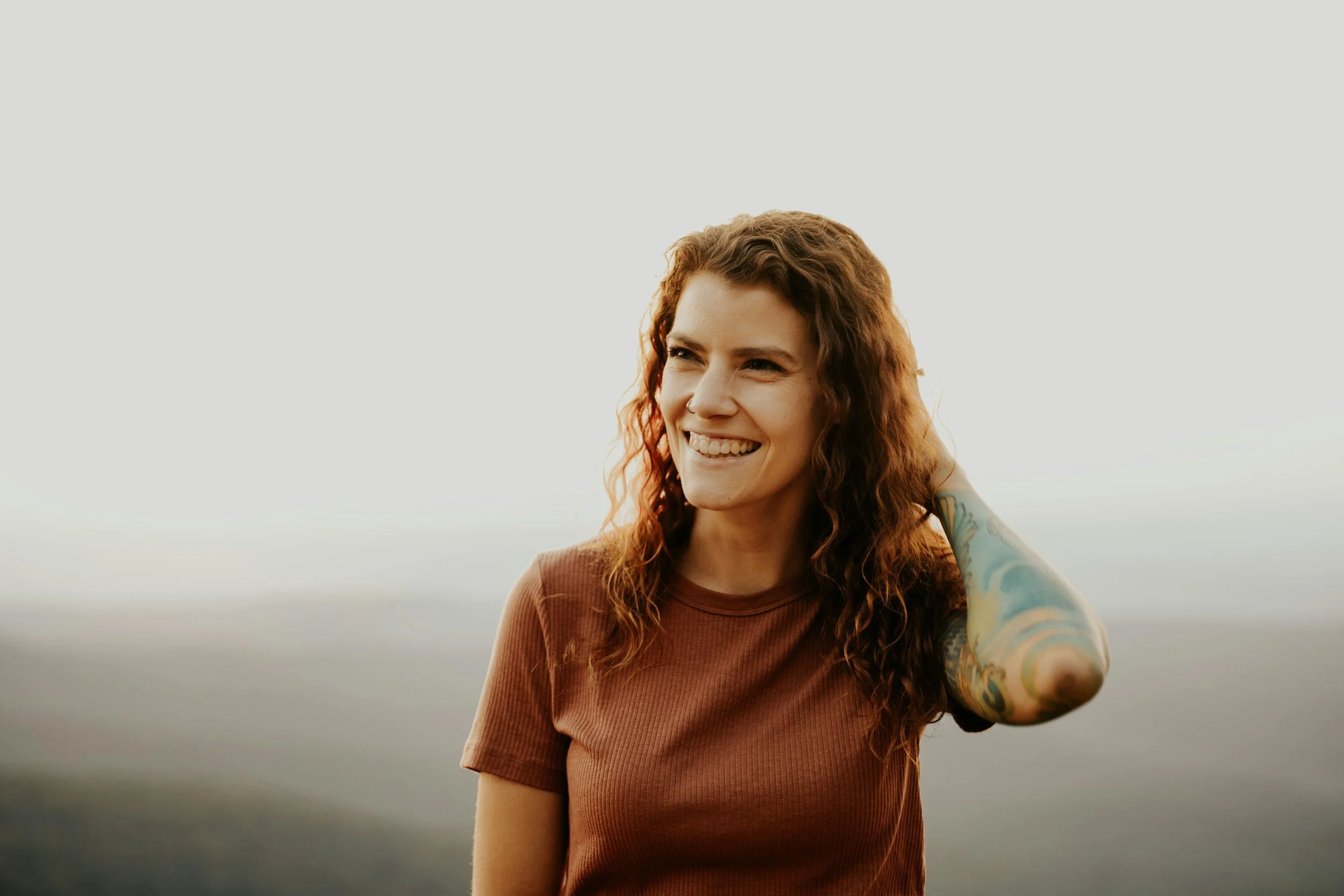 Woman with curly brown hair stands on a mountain, smiling, her hand is running through her hair and she has tattoos along her arm, EMDR therapy Great Barrington
