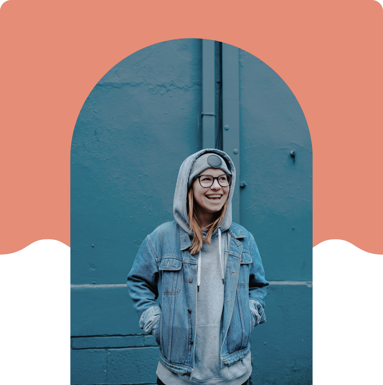 A young woman with glasses, wearing a gray beanie, hoodie, and denim jacket, smiling while standing against a blue wall.
