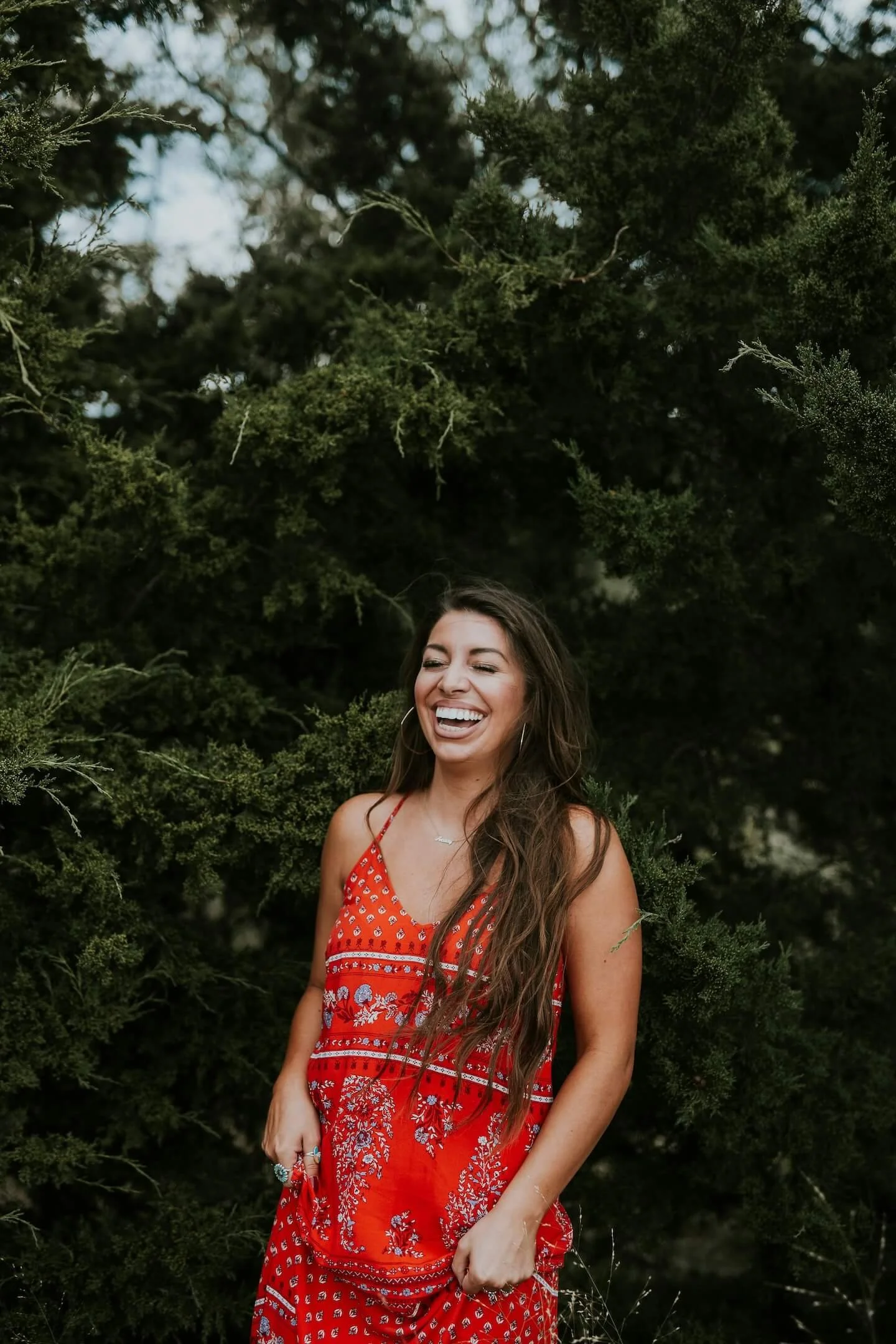 A woman laughing outdoors in a red patterned dress with lush green foliage in the background.