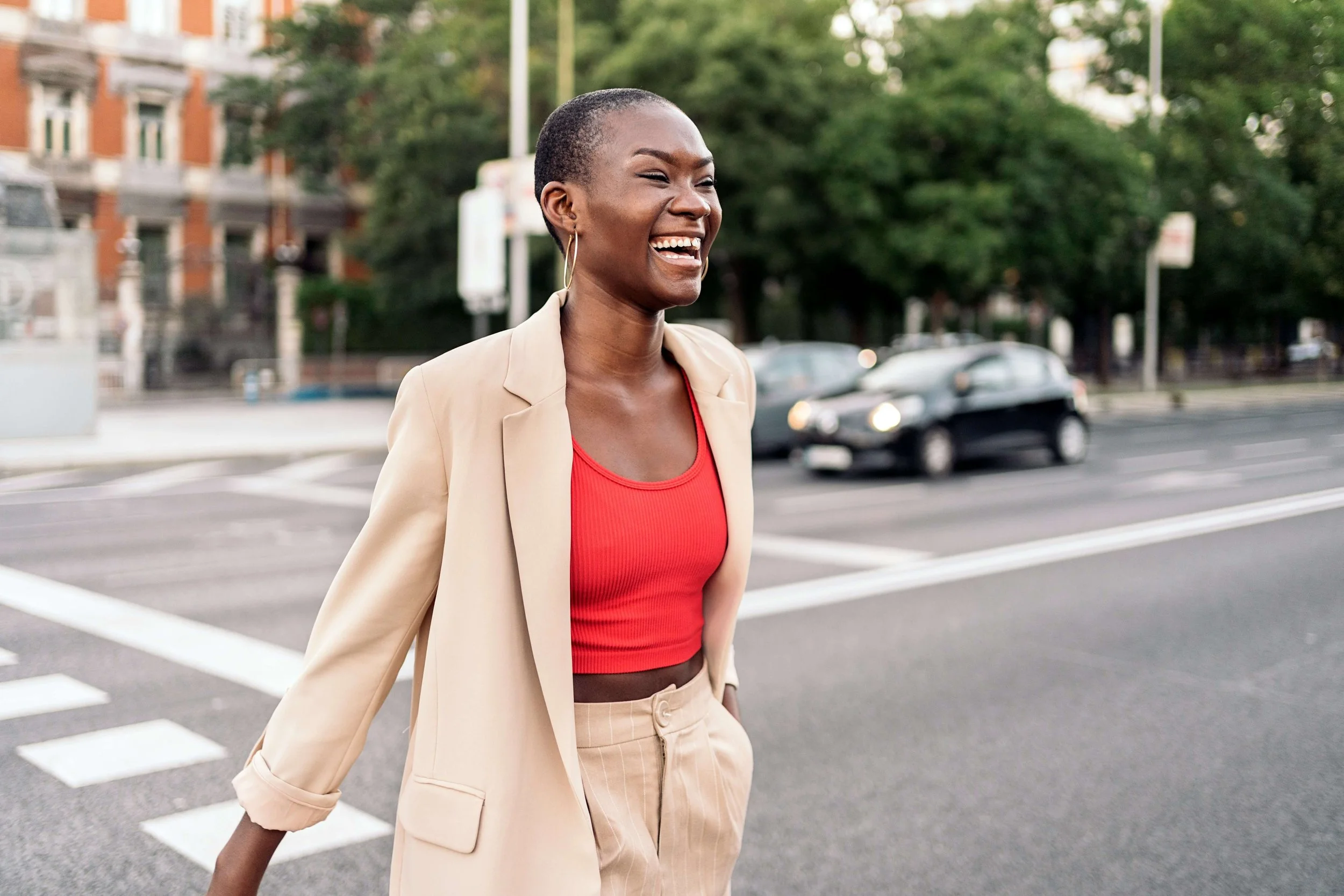 a Black woman with a buzz cut, wearing a bright, red top and khaki blazer, walks smiling across a city street
