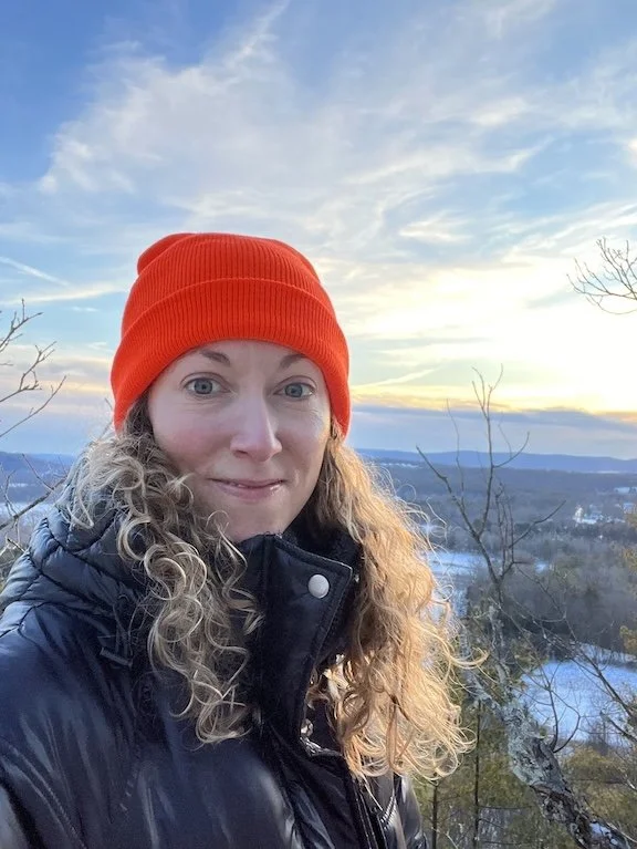 Woman with blonde, curly hair wearing an orange beanie and a black coat, stands on a mountain top, the sunset behind her, smiling softly at the camera, Rachel Duvall certified EMDR therapist MA FL