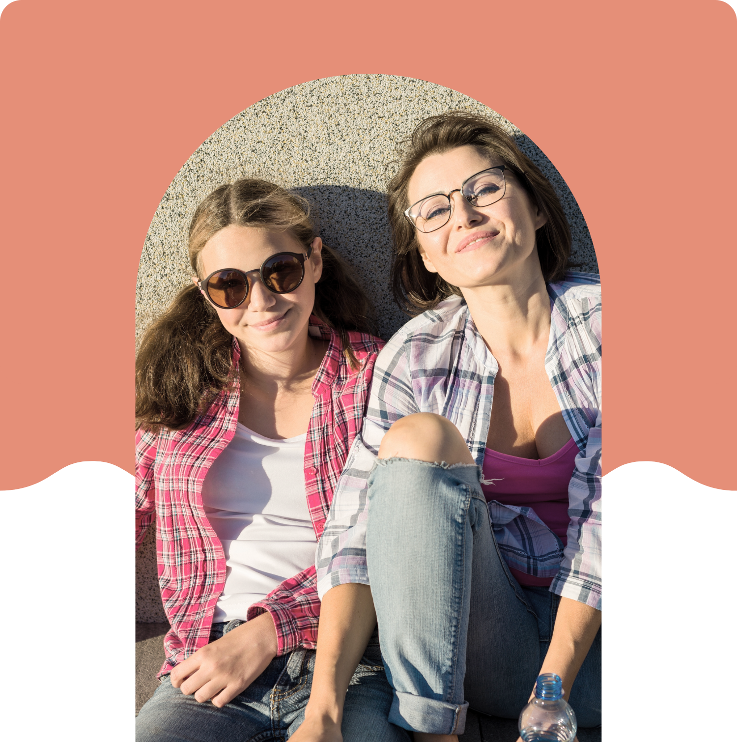 Two women sitting outdoors on a sunny day, smiling and looking at the camera. One woman is wearing sunglasses, a red plaid shirt, and white top, while the other woman is wearing glasses, a purple shirt, and a plaid shirt, with a water bottle beside her.