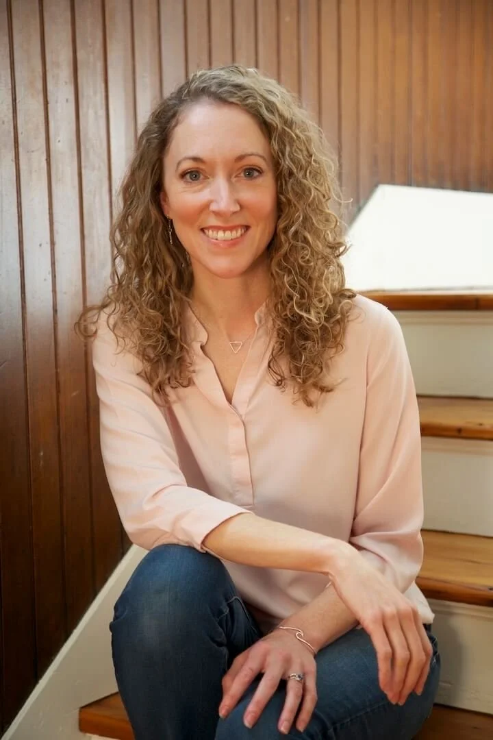White woman with curly blonde hair, wearing a pink button up shirt and jeans, sits on steps, smiling at the camera. Rachel Duvall EMDR and relational trauma therapist MA, NYC, and Boston
