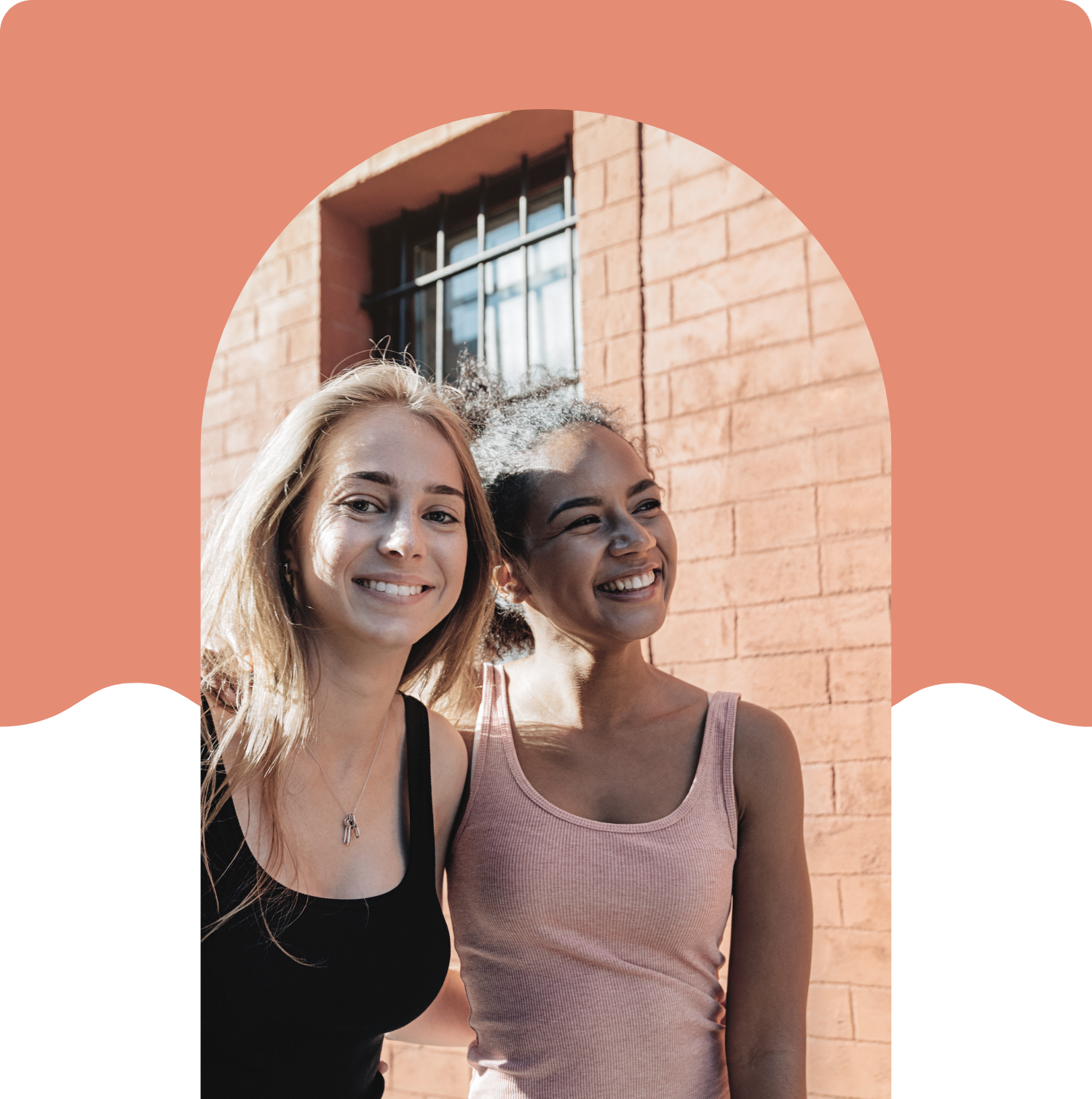 Two young women smiling and standing close outside in front of a brick wall with a window.