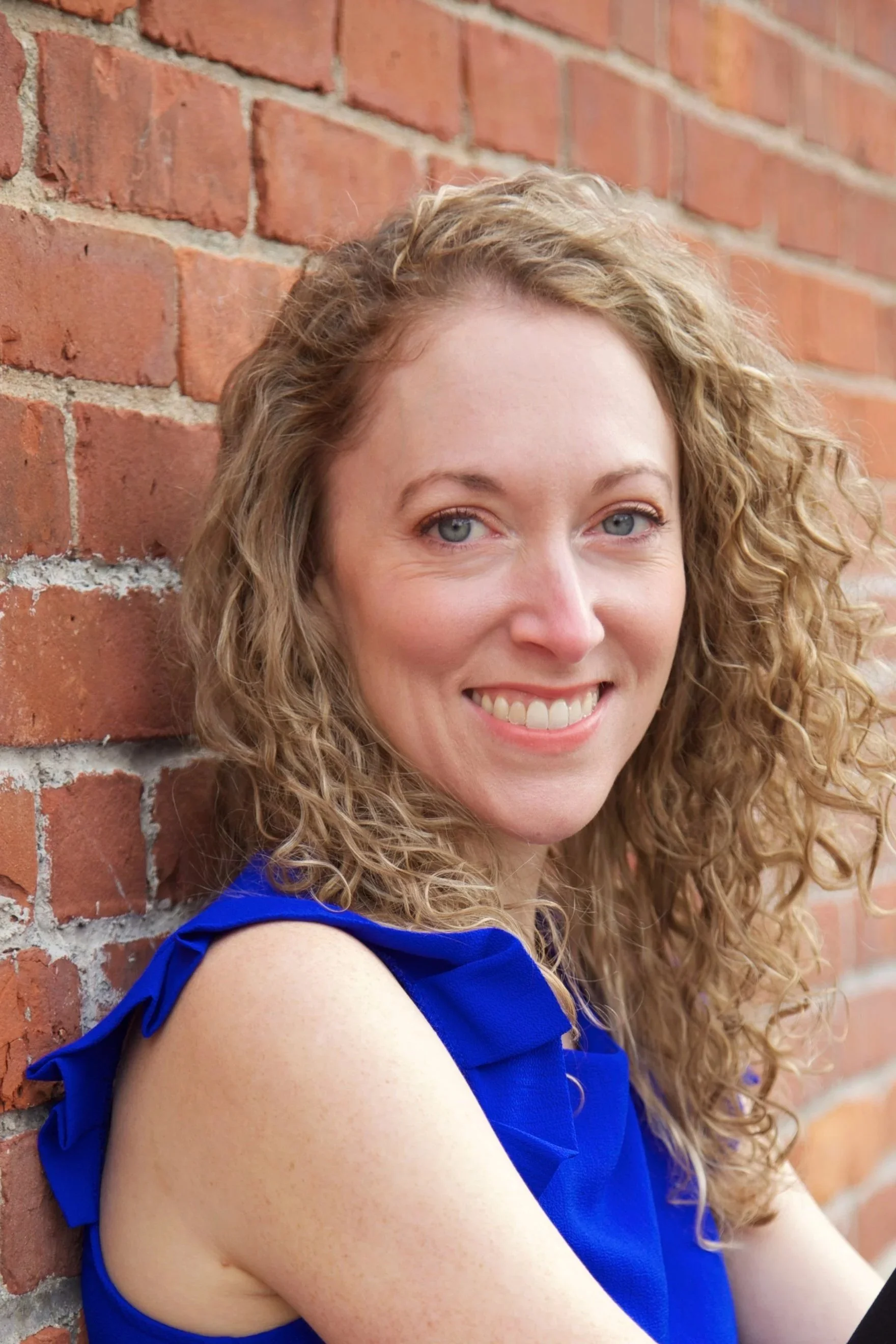White woman with blonde curly hair, wearing a blue sleeveless shirt, smiles while leaning on a brick wall, Rachel Duvall EMDR therapy intensives in the Berkshires near Boston and NYC