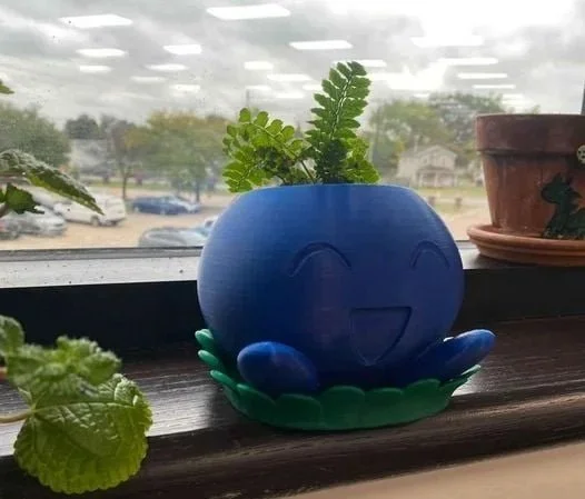 Blue smiling plant pot with green leaves inside, placed on a windowsill with other plants and a parking lot visible outside.