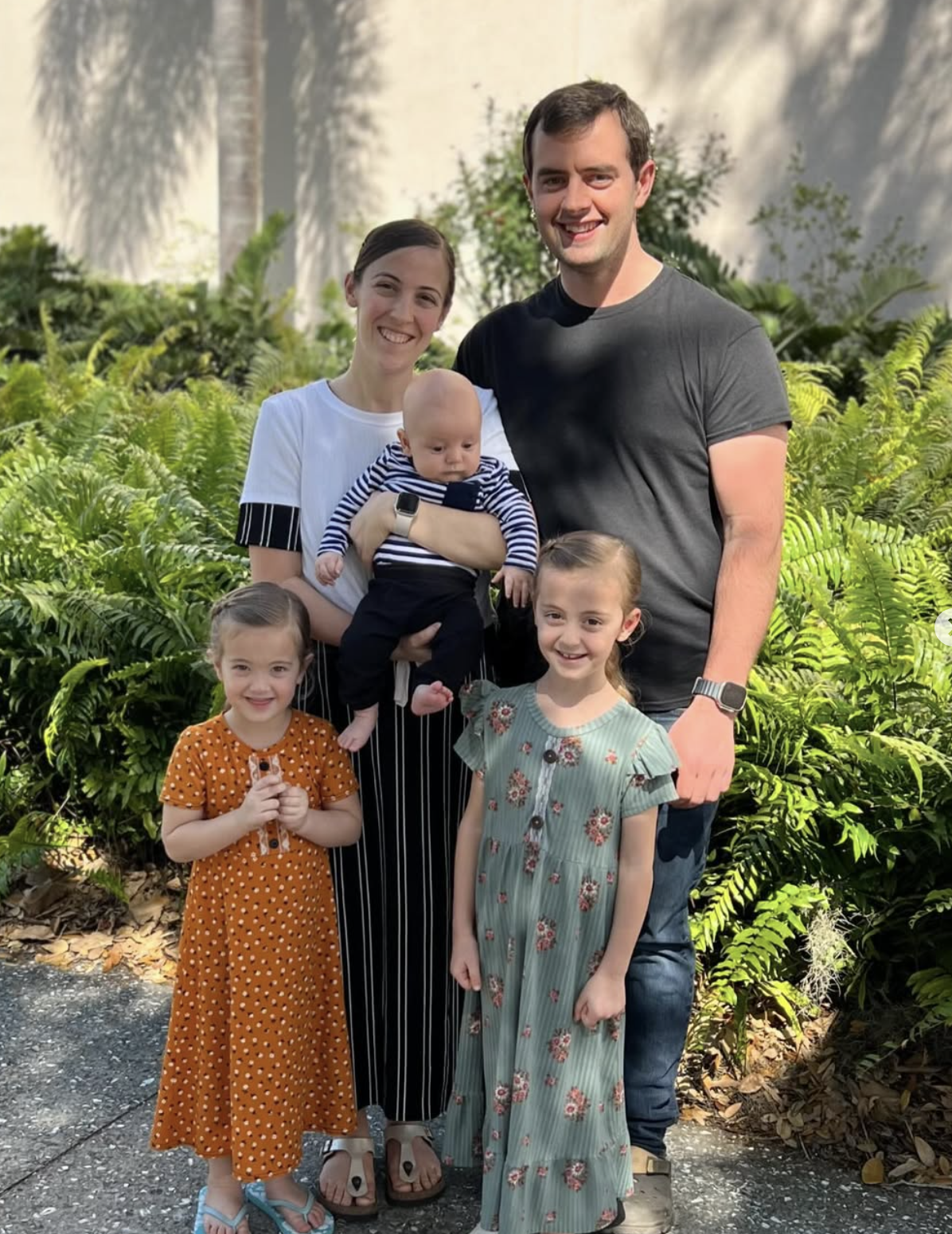 A family of six posing outdoors, with lush green plants and a white wall in the background. The mother is holding a baby, and two young girls are standing in front of the parents, smiling at the camera.